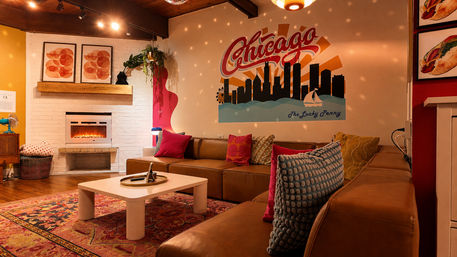 Cozy retro lounge with brown leather sectional, colorful throw pillows, low white coffee table, glowing electric fireplace and a large Chicago skyline mural under warm, dotted ambient lighting
