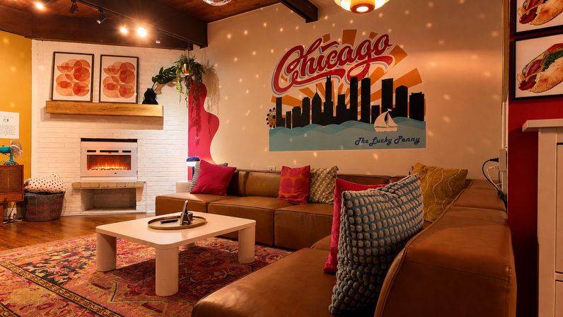 Cozy retro lounge with brown leather sectional, colorful throw pillows, low white coffee table, glowing electric fireplace and a large Chicago skyline mural under warm, dotted ambient lighting