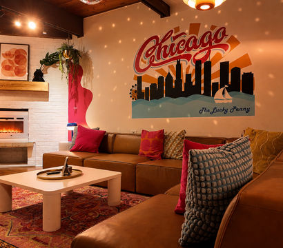 Cozy retro lounge with brown leather sectional, colorful throw pillows, low white coffee table, glowing electric fireplace and a large Chicago skyline mural under warm, dotted ambient lighting