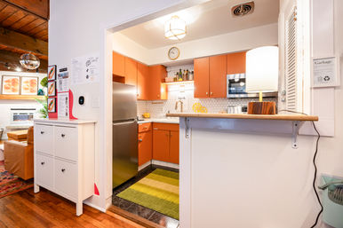 Cozy open-plan kitchen with retro orange cabinets, stainless steel refrigerator, hex tile backsplash, wood breakfast bar and lamp, living room peek.