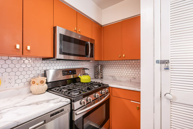 Cheerful modern kitchen with orange cabinets, stainless steel microwave and gas range, white hexagonal tile backsplash and marble-look countertops