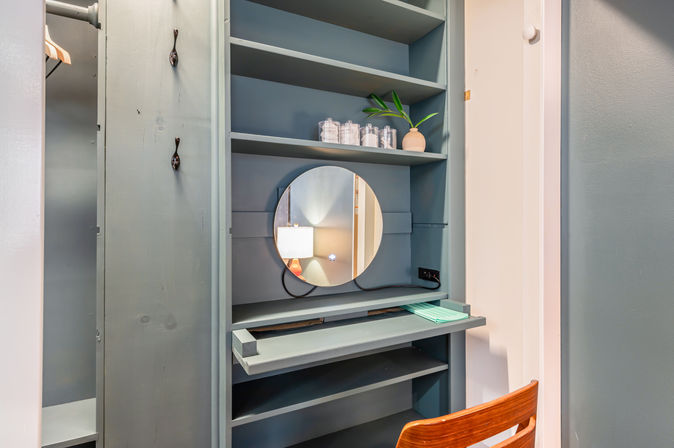Built-in teal closet vanity nook with round mirror, glass jars, small potted plant, pull-out shelf and wooden chair