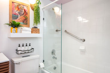 Modern white-tiled bathroom with glass shower door and bathtub, stainless grab bar, white toilet, floating shelf with folded towels, woven basket and hanging plant, playful framed tiger print and wall-mounted soap dispensers.