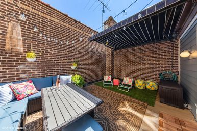 Cozy urban rooftop patio with brick walls, blue cushioned corner sofa and outdoor dining table, string lights and hanging planters, retractable awning, small artificial-grass play area with colorful folding chairs and patterned poufs.