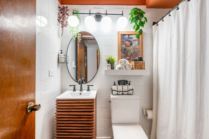 Bright, plant-accented white-tiled bathroom with wooden slatted vanity, oval mirror, black fixtures, floating shelf with rolled towels and basket, toilet with mounted dispensers, and white shower curtain.