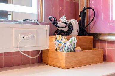 Pink-tiled vanity counter with wooden organizer holding colorful individually wrapped toothbrushes and toiletries, neatly coiled hairdryer and curling iron, and a plugged-in outlet beneath an illuminated mirror.