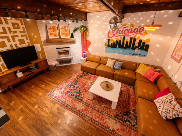 Cozy retro living room with caramel leather sectional, vibrant patterned rug, white coffee table, electric fireplace, mid-century TV console and hanging disco ball beneath a colorful Chicago skyline mural.
