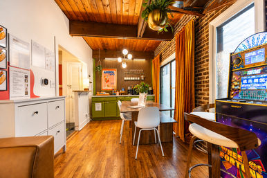 Cozy mid-century style dining nook with wood ceiling and floors, green kitchenette and Aperol poster, long wooden table with white chairs, exposed brick wall and orange velvet curtains, and a colorful arcade machine by the window.