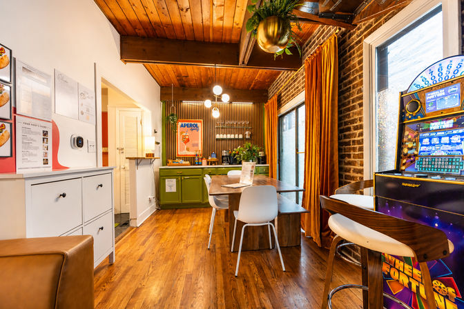 Cozy mid-century style dining nook with wood ceiling and floors, green kitchenette and Aperol poster, long wooden table with white chairs, exposed brick wall and orange velvet curtains, and a colorful arcade machine by the window.