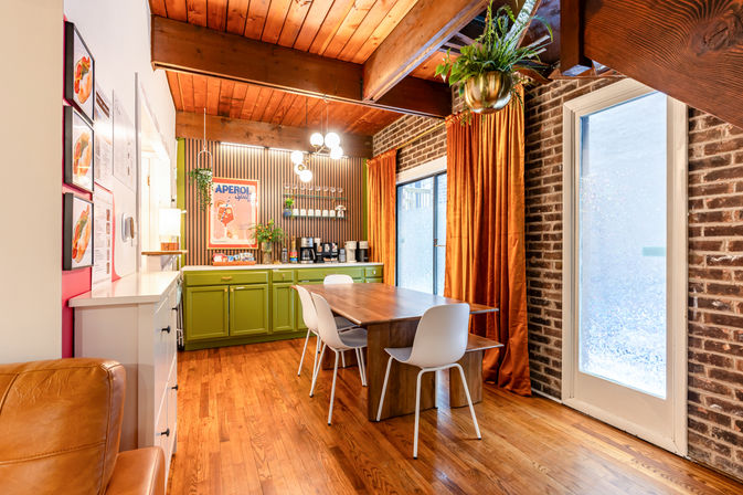 Cozy mid-century dining nook with exposed wooden beams and brick wall, green lower cabinets and coffee station, wooden dining table with white chairs, orange curtains and hanging plants.