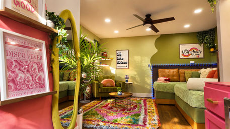 Vibrant disco-inspired living room with green walls, olive velvet armchair, tufted leather daybed trimmed in blue string lights, colorful shag rug, potted plants, gold oval mirror with small disco balls, and framed 'To The Disco' art.