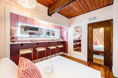 Loft-style bedroom with exposed wooden beams, pink tiled vanity wall and LED-lit mirrors above a maroon counter with gold-frame bar stools, white bed with textured coral throw pillow and rolled towel