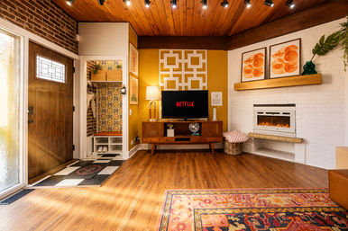 Cozy sunlit mid-century modern living room with wood ceiling, patterned area rug, fireplace, and vintage TV console displaying Netflix; bright entryway with wooden front door and retro wallpapered cubby.