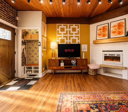 Cozy sunlit mid-century modern living room with wood ceiling, patterned area rug, fireplace, and vintage TV console displaying Netflix; bright entryway with wooden front door and retro wallpapered cubby.
