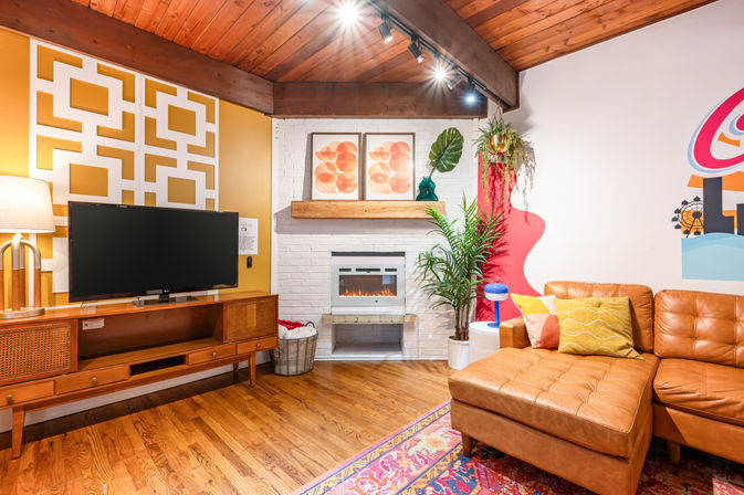 Cozy retro-modern living room with leather sectional chaise, wood-beamed ceiling, white brick fireplace and wooden mantel, TV on mid-century console, geometric yellow accent wall, colorful rug and houseplants.