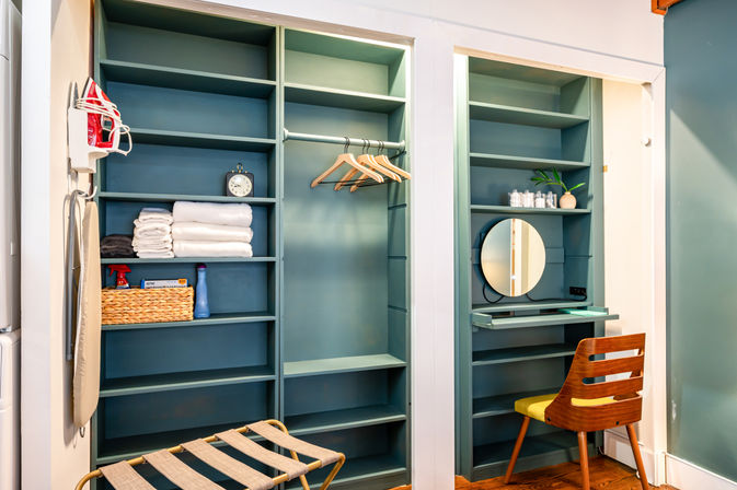 Cozy home dressing nook with built-in teal shelves, hanging rod with wooden hangers, stacked white towels, woven basket, wall-mounted iron, round vanity mirror and mid-century wooden chair on hardwood floor.