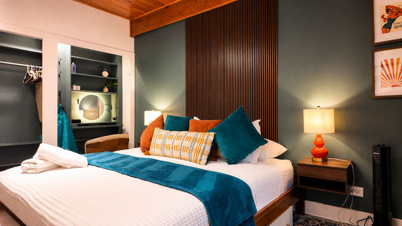Cozy modern bedroom with queen bed, teal throw and pillows, vertical wood-slat headboard, orange bedside lamp, open closet and round vanity mirror.