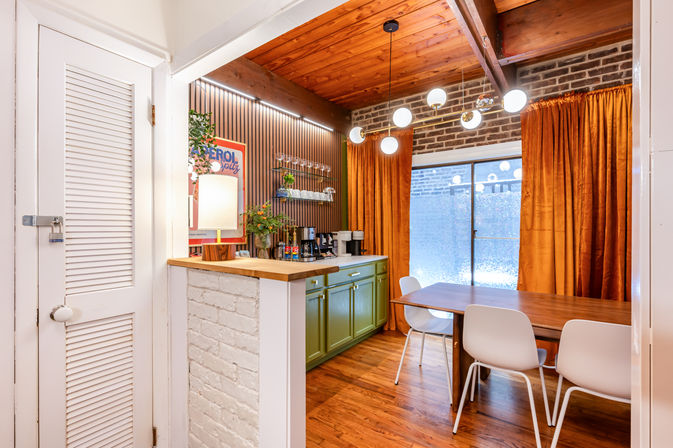 Cozy mid-century modern dining nook with wood-plank ceiling and exposed brick, green lower cabinets and wood countertop kitchenette, globe pendant lights, wooden dining table with white chairs, and rust-orange curtains over a sliding glass door.