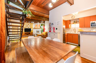 Cozy open-plan loft dining and living area with exposed wooden beams, large wooden dining table and white chairs, floating staircase with hanging plant, retro orange kitchen cabinets and hex-tile backsplash, and a comfy sofa by a fireplace.