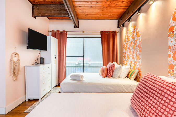 Cozy boho loft bedroom in a vacation rental with exposed wooden beams, two white-made beds with coral and patterned throw pillows, orange floral arched accent walls, pink curtains, wall-mounted TV and sliding glass door to a small balcony.