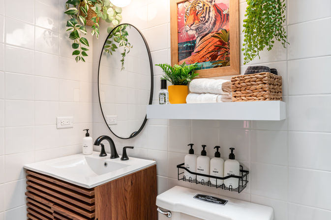 Bright modern small bathroom with white tiled walls, oval mirror over a wooden vanity with black faucet, hanging and potted greenery, bold tiger artwork, a floating shelf with rolled towels and wicker basket, and a wire rack of soap dispensers above the toilet.