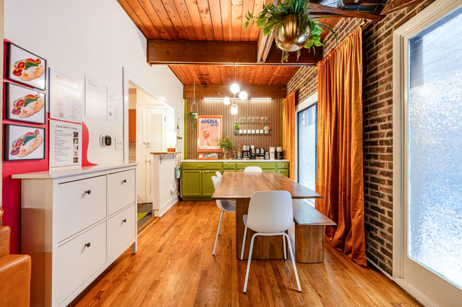 Cozy urban loft dining area with a long wooden table, white chairs and bench, green buffet coffee station, exposed brick wall, wood-plank ceiling, orange velvet curtains, hanging plant and pendant lights on warm hardwood floors.