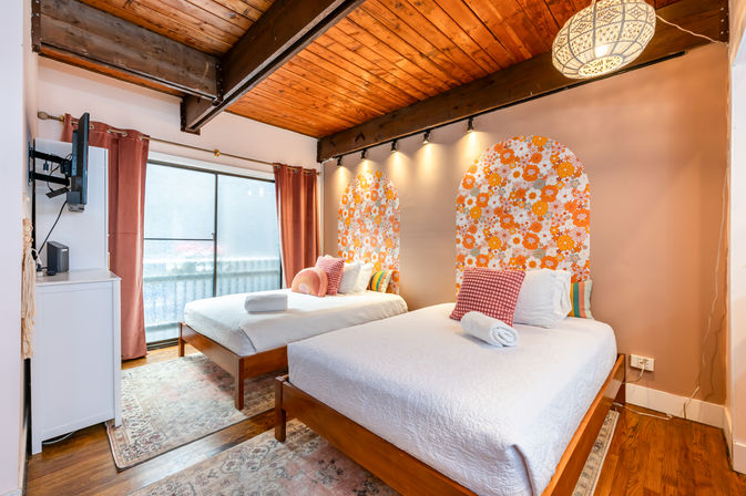 Cozy sunlit loft bedroom with exposed wooden beams, two twin beds with white linens and orange floral arched headboards, sliding glass door, patterned rugs and warm boho accents