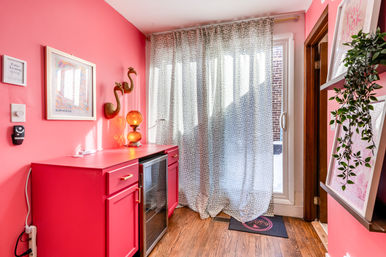 Vibrant pink entryway with a red cabinet and mini fridge, sheer patterned curtains over a sliding glass door, wood floor, gold flamingo wall decor, amber lamp and trailing plant.