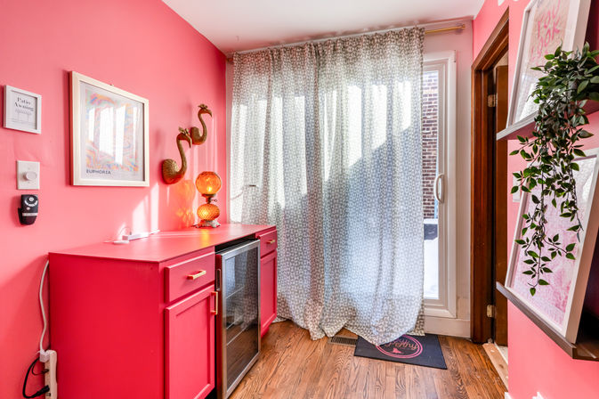 Vibrant pink entryway with a red cabinet and mini fridge, sheer patterned curtains over a sliding glass door, wood floor, gold flamingo wall decor, amber lamp and trailing plant.
