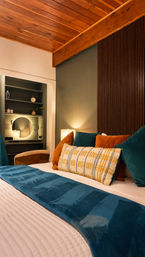 Cozy mid-century modern bedroom interior with warm wood-plank ceiling, teal bed runner, rust and teal velvet pillows, patterned lumbar cushion, bedside lamp, and built-in vanity with round mirror.