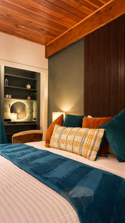 Cozy mid-century modern bedroom interior with warm wood-plank ceiling, teal bed runner, rust and teal velvet pillows, patterned lumbar cushion, bedside lamp, and built-in vanity with round mirror.