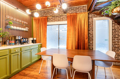 Cozy mid-century modern dining room with olive-green cabinets and a coffee station against an exposed brick wall, wooden dining table with white chairs, orange velvet curtains and globe pendant lighting.