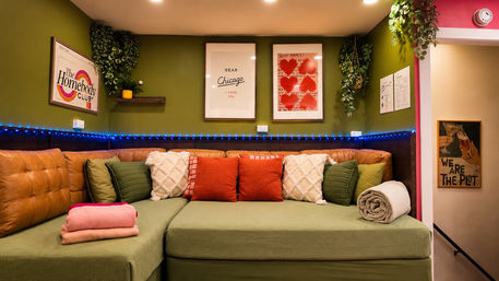 Cozy Chicago‑themed lounge nook with green sectional, tan leather back cushions, red and cream throw pillows, hanging plants, framed heart art and blue LED string lights.