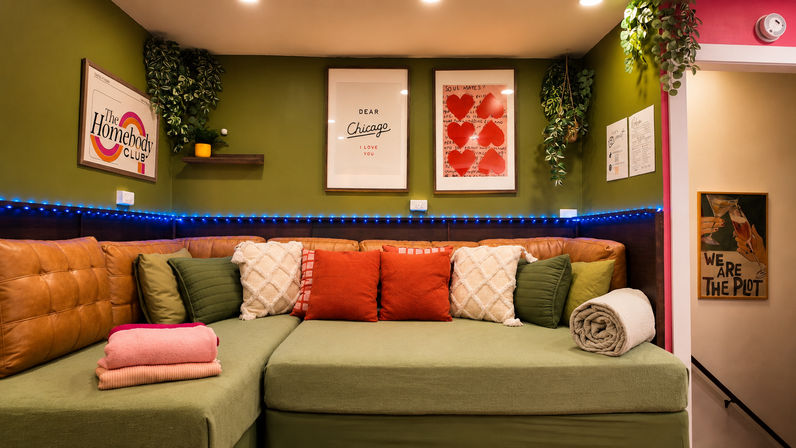 Cozy Chicago‑themed lounge nook with green sectional, tan leather back cushions, red and cream throw pillows, hanging plants, framed heart art and blue LED string lights.