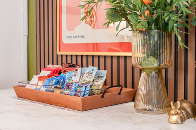 Cheerful snack display: wooden tray of assorted packaged snacks on a marble countertop beside a tall ribbed glass vase with leafy branches and oranges, colorful framed art on a wood‑slat wall.