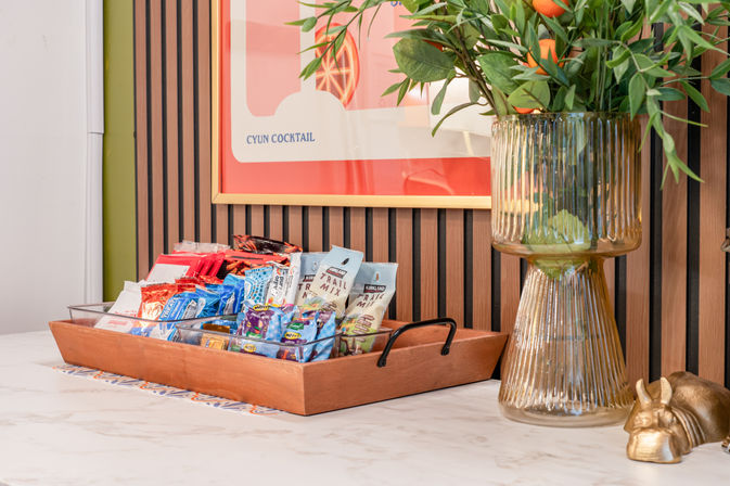 Cheerful snack display: wooden tray of assorted packaged snacks on a marble countertop beside a tall ribbed glass vase with leafy branches and oranges, colorful framed art on a wood‑slat wall.