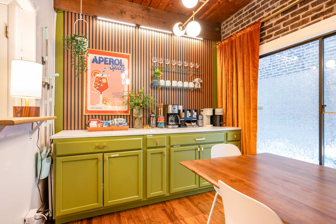 Cozy mid-century modern dining nook with olive-green cabinets and white countertop coffee bar, striped wood accent wall, Aperol Spritz poster, hanging plant, orange velvet curtains, and wooden dining table.