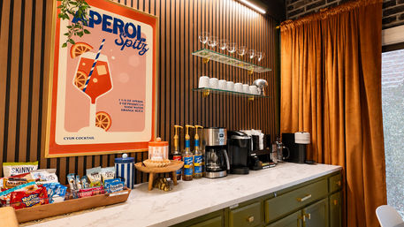 Cozy modern coffee station interior with Aperol Spritz poster, marble countertop, espresso machines, syrup bottles, glass mugs on floating shelves, wood‑slat wall and rust-colored curtains.