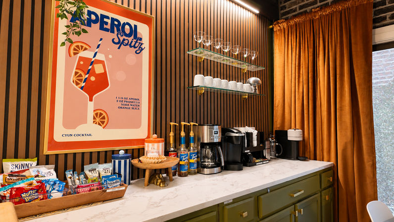 Cozy modern coffee station interior with Aperol Spritz poster, marble countertop, espresso machines, syrup bottles, glass mugs on floating shelves, wood‑slat wall and rust-colored curtains.