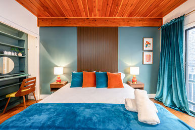 Cozy modern bedroom with teal accent wall and velvet curtains, warm wood ceiling, bed with teal and orange pillows, bedside lamps and a small desk