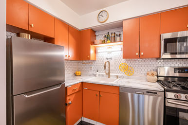 Retro-inspired kitchen with bright orange cabinets, stainless steel fridge, dishwasher, gas range and microwave, hex-tile backsplash, marble countertop and gold-tone faucet.