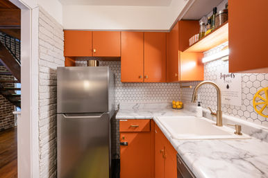 Cozy urban kitchen with terra cotta orange cabinets, stainless steel fridge, white marble countertops, hexagon tile backsplash, brass faucet over a farmhouse sink and a pop of lemons