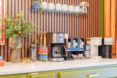 Cozy countertop coffee station with stainless drip coffee maker, two white single-serve brewers, syrup pumps, French press, mugs on a glass shelf, and a vase of orange branches against a vertical wood-slat wall.