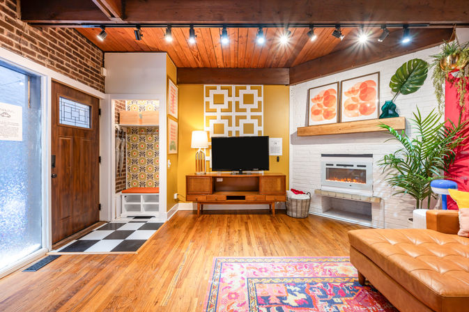 Cozy mid-century modern living room with wood floors, checkerboard entry, mustard accent wall, TV console, electric fireplace, colorful rug, and indoor plants.
