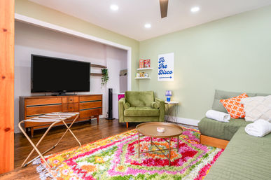 Playful retro-chic living room with flat-screen TV on mid-century wood console, green armchair and sectional, colorful shag rug, round coffee table, 'To The Disco' wall art and hardwood floors.