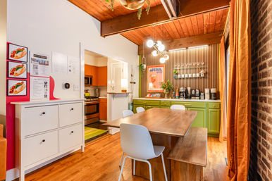 Cozy urban apartment dining area with a wooden table and bench, white chairs beneath exposed wood-beam ceiling and globe chandelier, green lower cabinets and coffee station on a slatted wood accent wall, exposed brick wall and orange curtains, view into a compact stainless-steel kitchen.