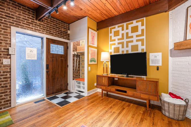Cozy mid-century modern entry and living area with wooden front door and frosted sidelight, mustard yellow accent wall with geometric panel, retro wood TV console and flat-screen, checkerboard tile entry by a bench with hooks, exposed brick and warm hardwood floors.
