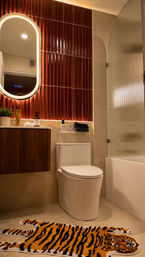 Contemporary bathroom interior with glossy vertical red tiles, backlit oval mirror above a wood vanity, white toilet, ribbed glass shower door, and a playful tiger-striped bath rug.