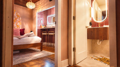 Cozy pink boutique guest room with wooden bed, floral arched headboard, textured pillows and folded towels, opening into an en-suite bathroom with illuminated oval mirror, red tile, brass fixtures, two bar stools at a vanity and a tiger-print bath mat, warm ambient lighting.