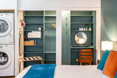 Cozy modern bedroom with stacked washer-dryer, open teal built-in shelving and closet, vanity nook with round mirror and wooden chair, and a bed dressed in white linen with blue throw and orange and teal pillows.